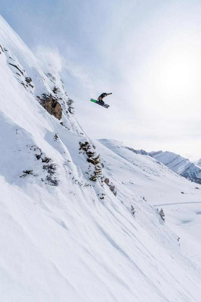 Skier launches a cliff into powder on a steep alpine face under a hazy sun.