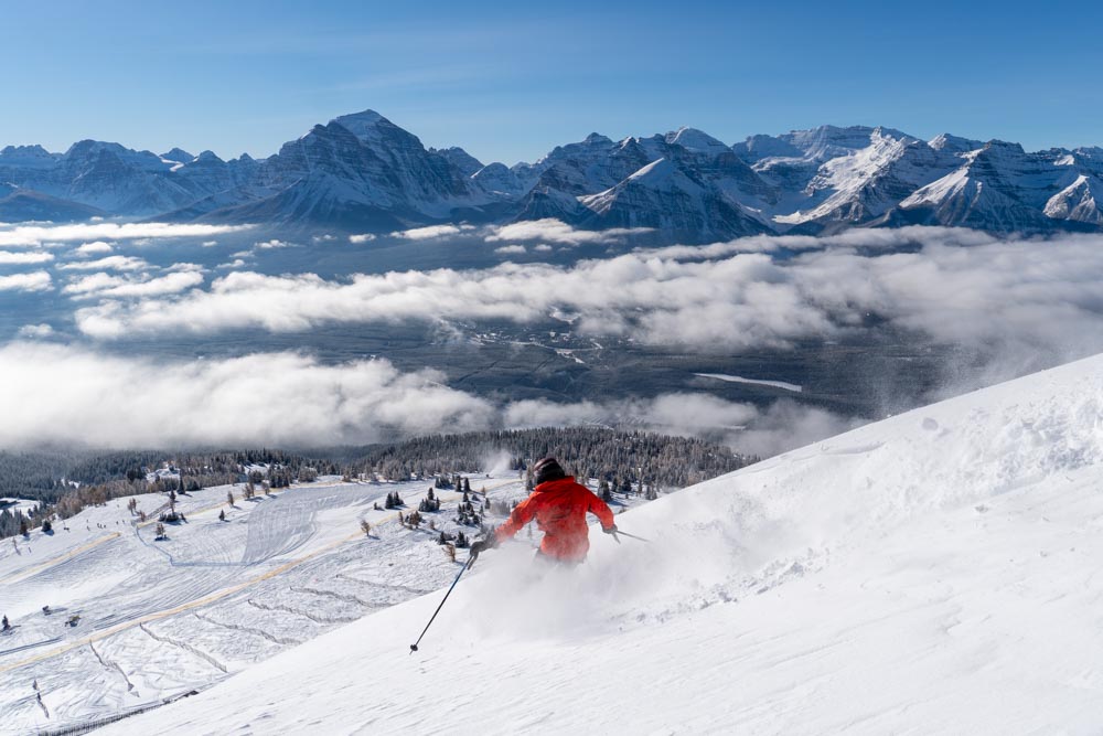 Skier in a red jacket arcs turns above a sea of clouds with Rocky Mountain peaks beyond.