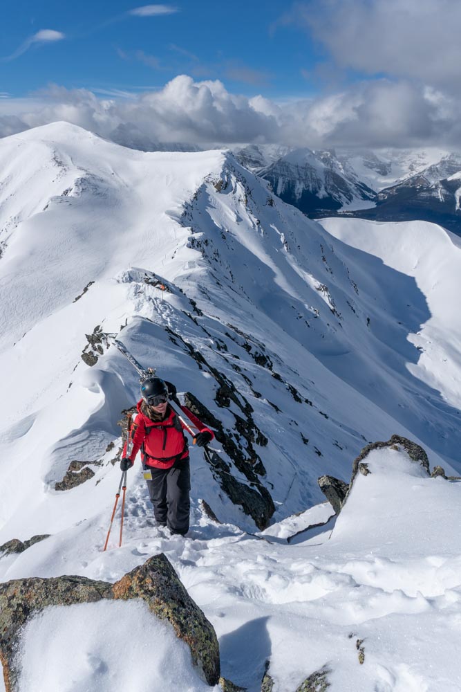 Skier bootpacks along a narrow, snow‑covered alpine ridge under a blue sky.