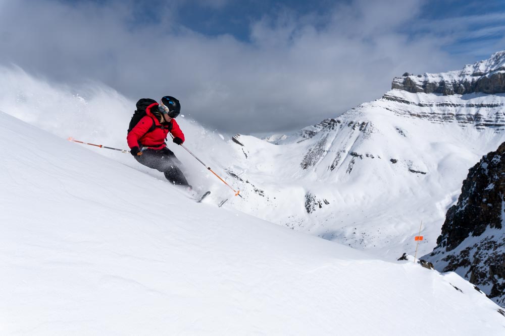 Skier in a red jacket carves a steep powder slope as spindrift trails across an upper‑mountain bowl.