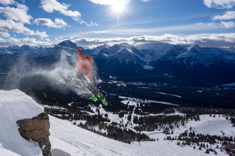 Skier explodes a cliff drop with spray, Lake Louise valley and peaks behind.
