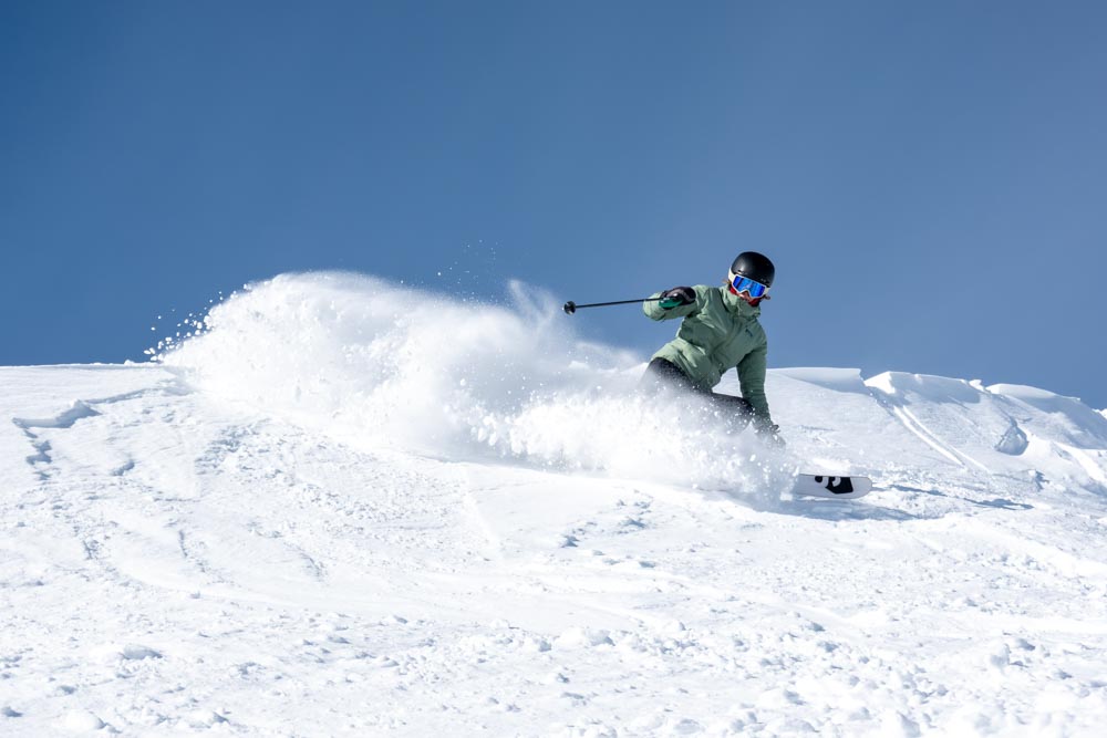 Original: Skier slashes a powder turn on a bright upper‑mountain bowl under a clear blue sky.