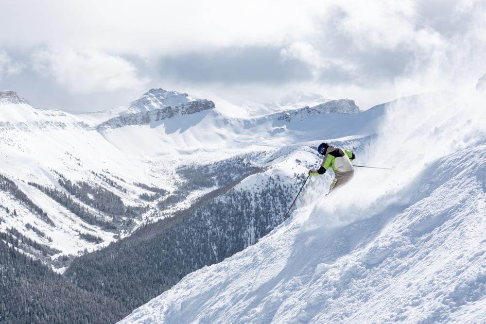 Skier drives a powerful turn down a steep upper‑mountain chute as a snow plume rises, Unity Peak beyond.