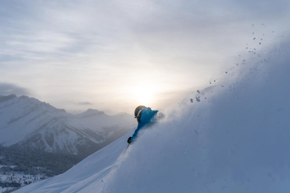 Skier carves a sunrise powder turn, snow plume backlit against alpine peaks.