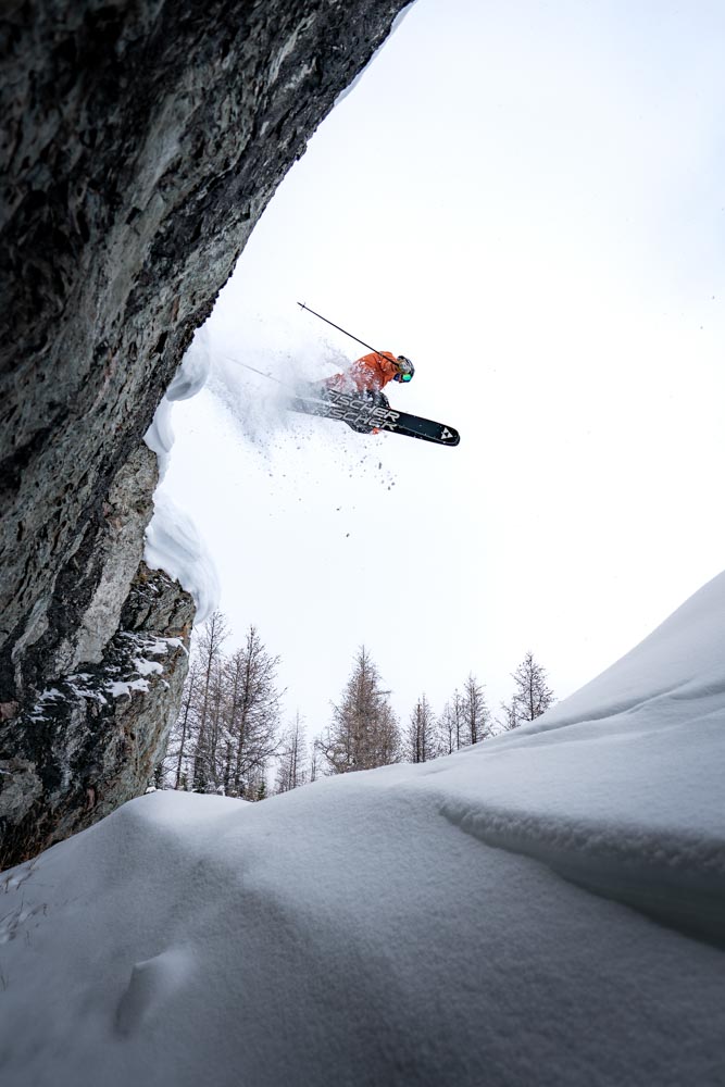 Skier launches off a rock face, throwing powder against an overcast sky with winter trees below.