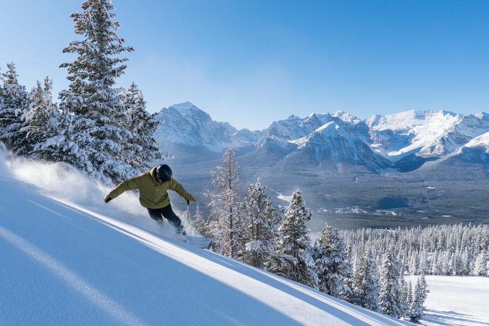 Original: Snowboarder carves fresh powder on a sunlit slope with snow‑covered trees and Rocky Mountain peaks in the background.