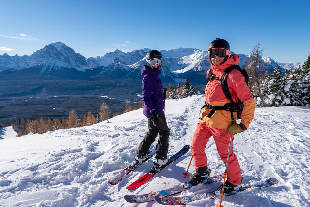Two skiers pause on a sunny ridge with panoramic Rocky Mountain peaks in the background.