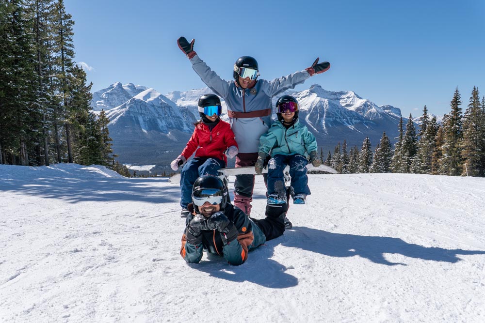 Family poses playfully on a groomed run with Rocky Mountain peaks in the background.