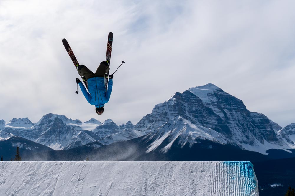Freestyle skier executes an inverted flip off a large jump with Rocky Mountain peaks in the background.
